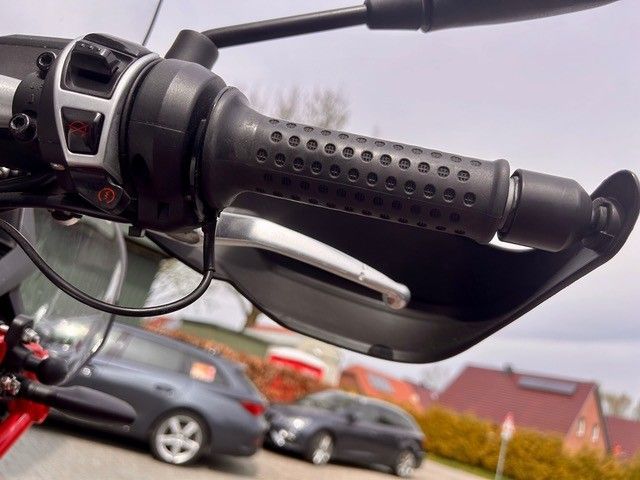 Nahaufnahme eines Motorradlenkers und Bremshebels; im Hintergrund sind geparkte Autos verschwommen zu sehen.