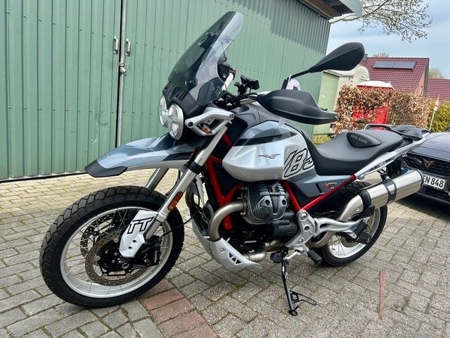 Silbernes Tourenmotorrad, geparkt neben einer grünen Mauer und Blumen, Seitenansicht auf dem Seitenständer.