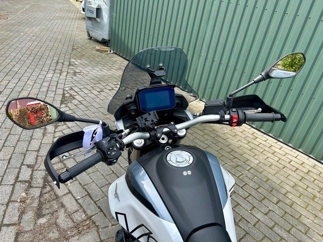 Motorradlenker und Armaturenbrett von oben gesehen, geparkt neben einer grünen Wellblechmauer und einem Backsteinpflaster.