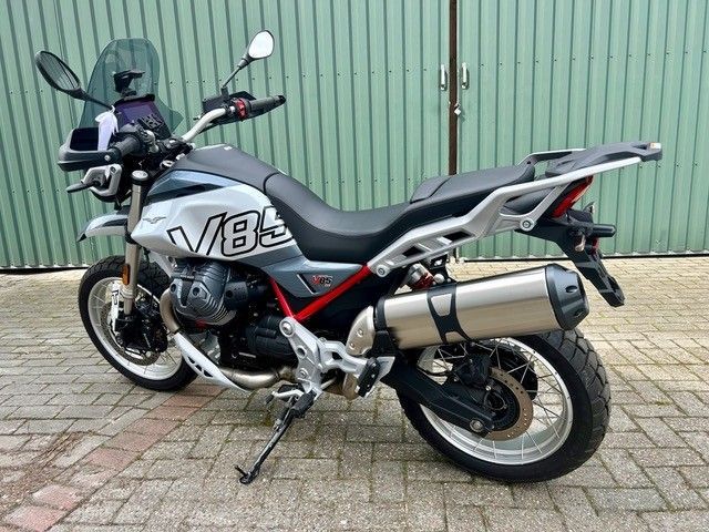 Ein silber-schwarzes Adventure-Motorrad parkt auf einem Backsteinpflaster vor einer grünen Wellblechwand.