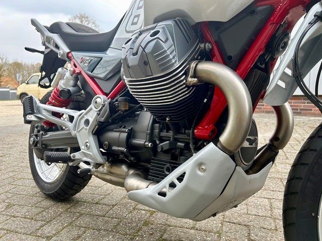 Nahaufnahme eines silbernen Motorradmotors mit Auspuff auf einem roten Rahmen, der auf einer gepflasterten Fläche abgestellt ist.