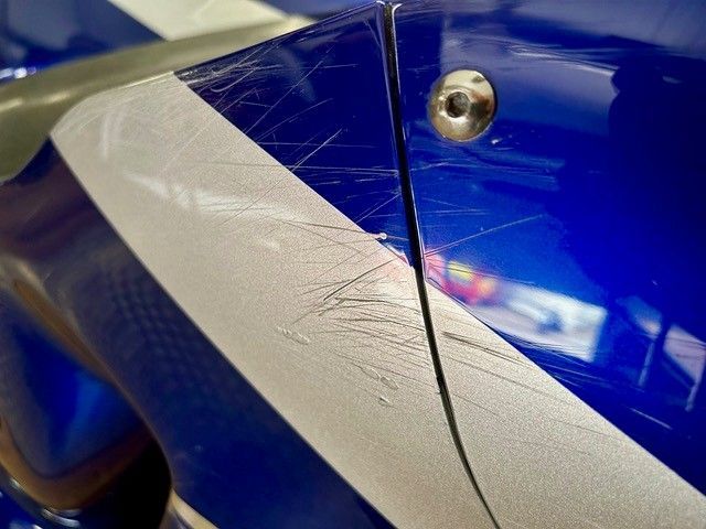 Nahaufnahme einer blauen Fahrzeugkarosserie mit einem weißen Diagonalstreifen und einem sichtbaren Riss in der Nähe eines Schraubenlochs