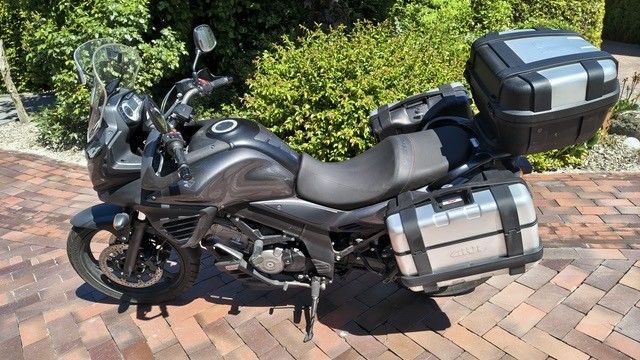 Schwarzes Tourenmotorrad mit Hartschalenkoffern und Topcase, geparkt auf einer gepflasterten Auffahrt in der Nähe von Hecken.
