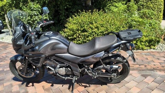 Ein schwarzes Tourenmotorrad parkt auf einer gepflasterten Auffahrt neben grünen Büschen und Bäumen.