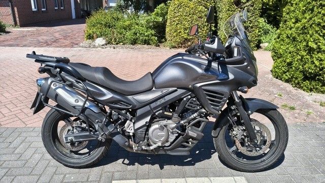 Schwarzes Tourenmotorrad parkt auf einer gepflasterten Auffahrt vor einem Haus und Sträuchern.