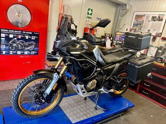 Schwarzes Motorrad auf einer blauen Hebebühne in einer Werkstatt, daneben rote Werkzeugschränke und Geräte.