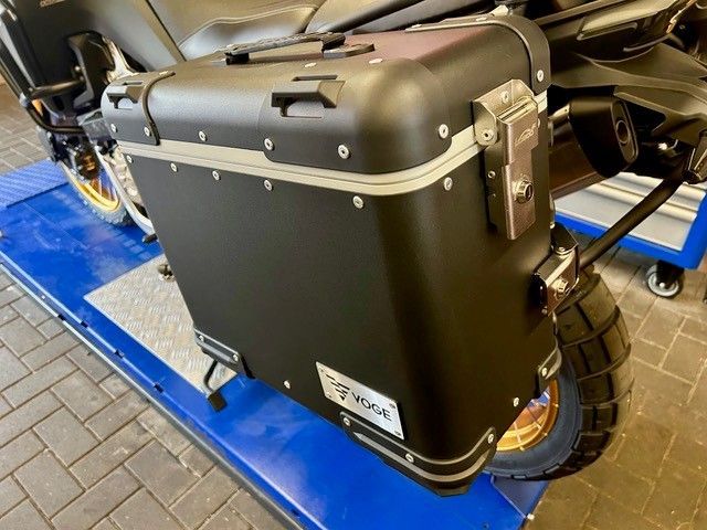 Schwarze, harte Motorradkoffer mit silbernen Zierelementen an einem Motorrad in einem Ausstellungsraum auf blauem Boden.
