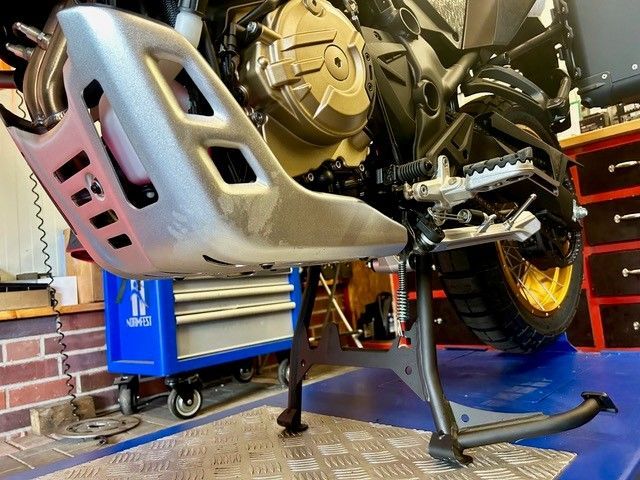 Motorrad auf einem Ständer in einer Garage, im Hintergrund ein silberner Auspuffschutz und eine blaue Werkzeugkiste.