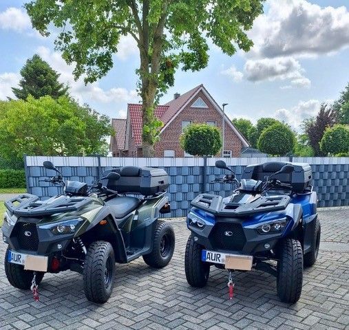 Zwei Geländefahrzeuge, eines grün und eines blau, parkten nebeneinander auf einer gepflasterten Fläche vor einem Haus mit Backsteinfassade.