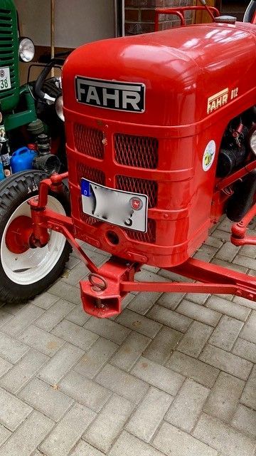 Roter Fahr-Traktor mit weißen Rädern, Nummernschild und Frontkupplung auf einem gepflasterten Platz.