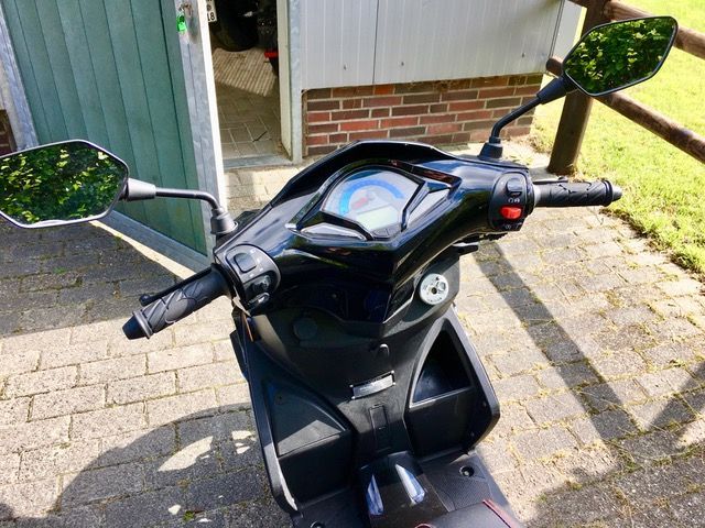 Ein schwarzer Motorroller ist auf einem gepflasterten Gehweg vor einem Backsteingebäude geparkt.