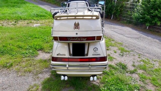 Rückansicht eines goldenen Honda Goldwing-Motorrads mit Gepäckträger und Doppelauspuff, auf Gras geparkt.