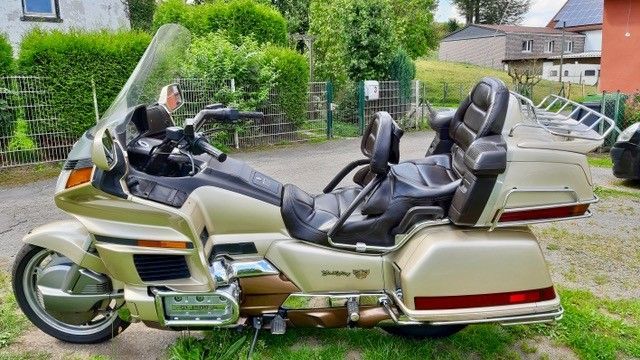 Goldenes Motorrad, eine Honda Goldwing, geparkt auf einem gepflasterten Platz. Es hat eine große Windschutzscheibe, bequeme Sitze und Gepäckfächer.