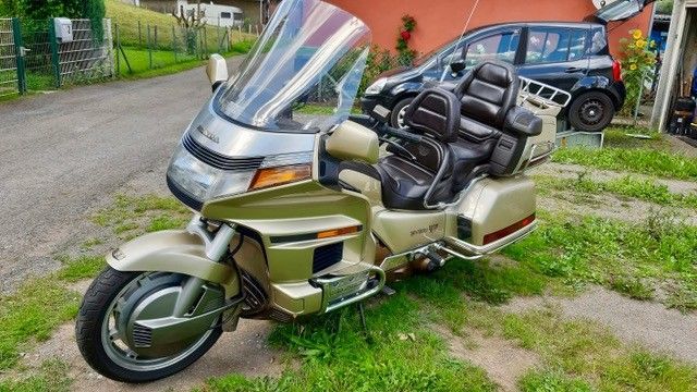 Ein goldenes Honda Goldwing-Motorrad, das auf Gras geparkt ist. Es hat eine große Windschutzscheibe und einen Beifahrersitz.