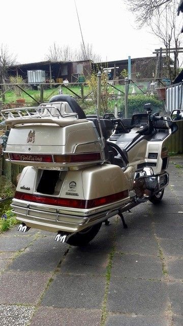 Gold- und braunes Honda Goldwing-Motorrad, das im Freien auf einer gepflasterten Fläche geparkt ist. Es hat Gepäckfächer und verchromte Auspuffrohre.