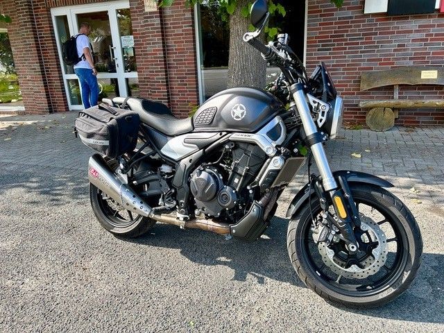 Ein dunkelgraues Motorrad parkt vor einem Backsteingebäude. Das Motorrad hat Satteltaschen und einen nachgerüsteten Auspuff.