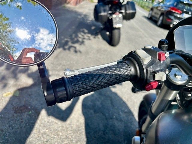 Nahaufnahme eines Motorradlenkers mit einem Spiegel, der einen sonnigen Himmel und die Hand des Fotografen reflektiert. Im Hintergrund sind andere Motorräder zu sehen.
