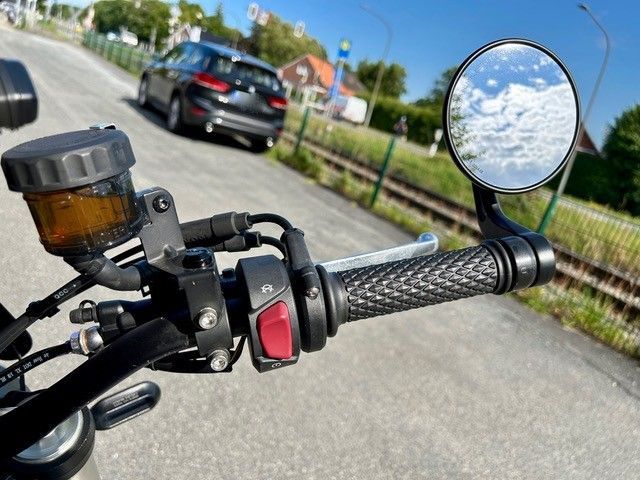 Motorradlenker mit einem Spiegel, der eine verschwommene Landschaft reflektiert. Das Motorrad steht auf einer Straße, im Hintergrund ist ein Auto zu sehen.