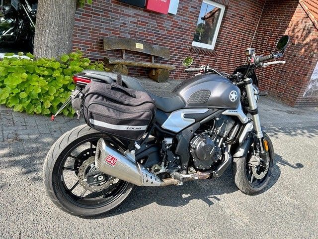 Ein graues CFMOTO-Motorrad mit schwarzen Satteltaschen, das vor einem Backsteingebäude geparkt ist. Es hat einen silbernen Auspuff und schwarze Räder.