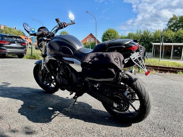 Ein dunkelgraues Motorrad parkt auf dem Bürgersteig neben einem Gebäude und einem Auto. Schwarze Satteltaschen und ein Nummernschildhalter sind sichtbar.