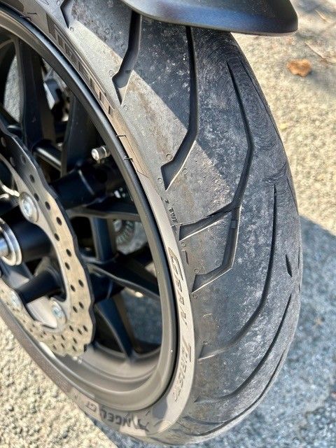 Nahaufnahme eines Motorrad-Hinterreifens mit sichtbarem Profil. Der Reifen ist schwarz und hat abgenutzte Kanten, eine schwarze Felge und eine sichtbare Scheibenbremse.