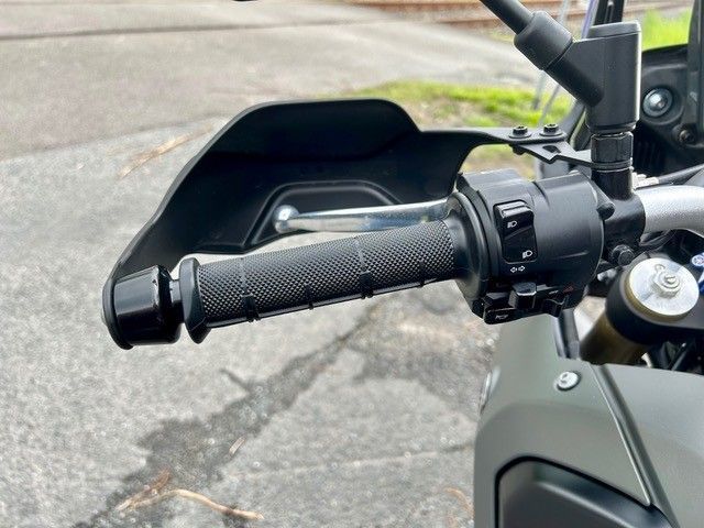 Nahaufnahme eines Motorradlenkers mit schwarzem Handschutz und strukturiertem Griff auf grauer Straße.