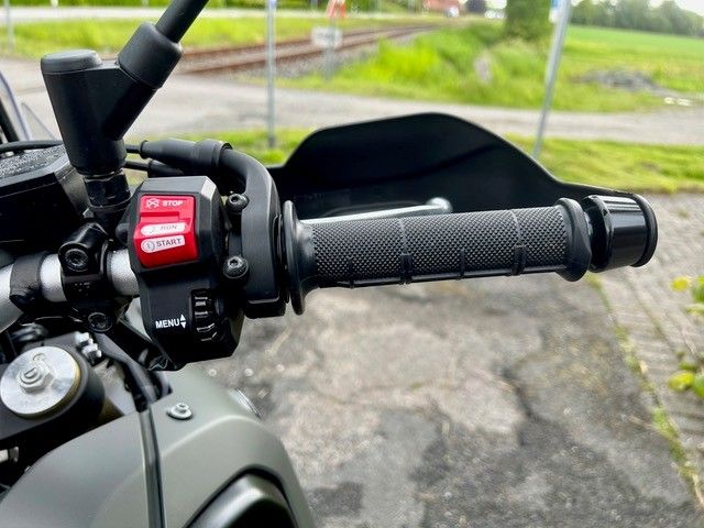 Nahaufnahme eines Motorradlenkers mit schwarzem Griff und rotem Schalter auf einer gepflasterten Straße.