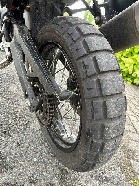 Nahaufnahme eines Motorradhinterreifens und -rads auf Schotter, die das Profil und den Kettenantrieb zeigt.