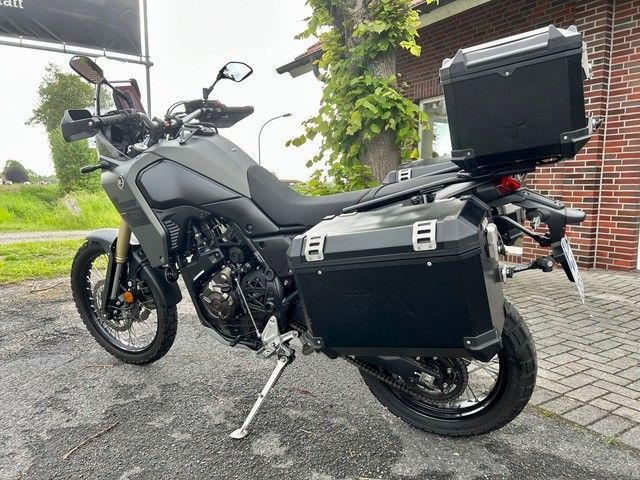 Schwarzes Adventure-Motorrad, mit Seitenkoffern und Topcase, parkt auf nassem Gehweg neben einer Backsteinmauer und Grünflächen.