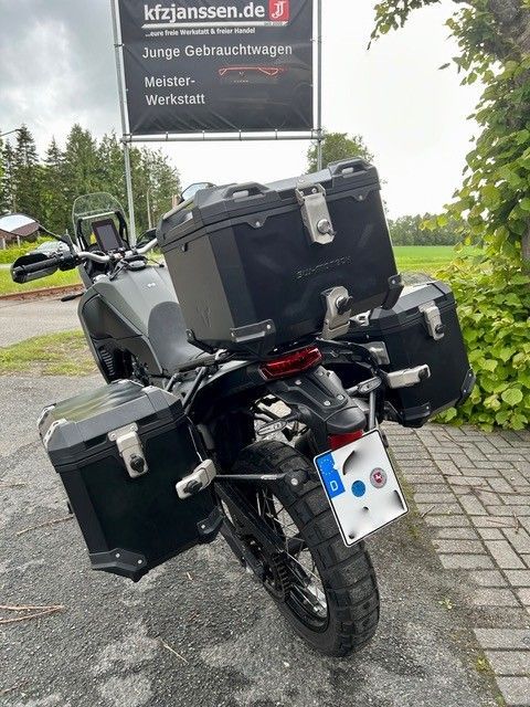 Schwarzes Tourenmotorrad mit schwarzen Seitenkoffern und Topcase, geparkt im Freien am nassen Straßenrand.