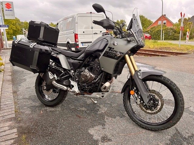 Graues Adventure-Motorrad mit schwarzen Seitenkoffern parkt auf einer nassen Straße in der Nähe von Häusern.