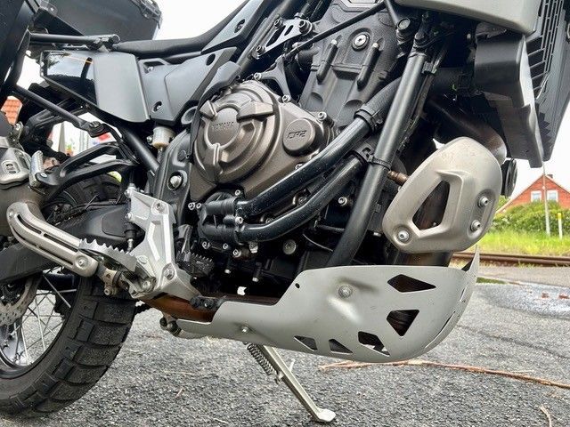 Nahaufnahme des Motors und des Unterfahrschutzes eines Motorrads am nassen Straßenrand.