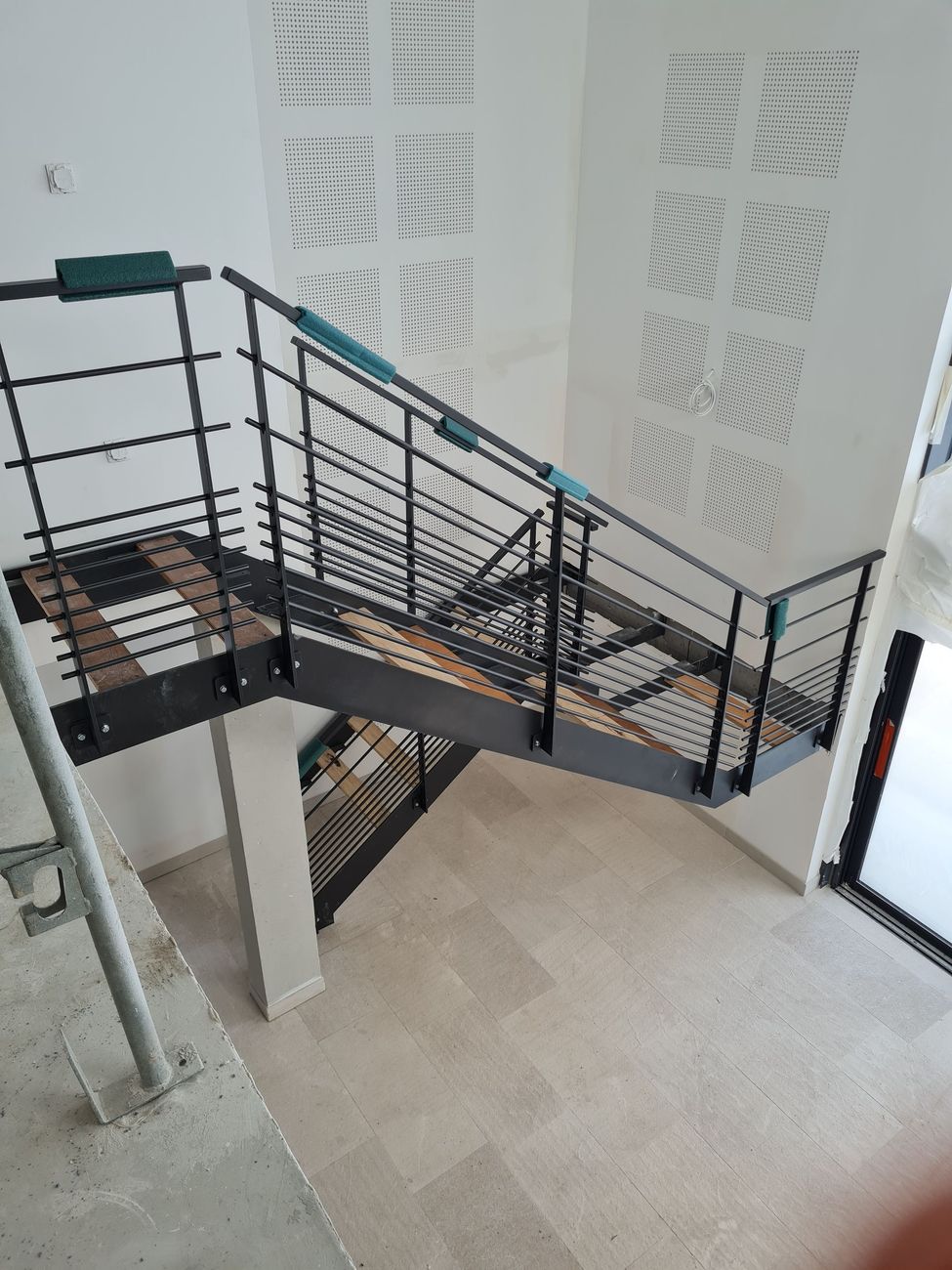 Escalier intérieur avec rampes noires et sol carrelé beige dans un bâtiment aux murs blancs.