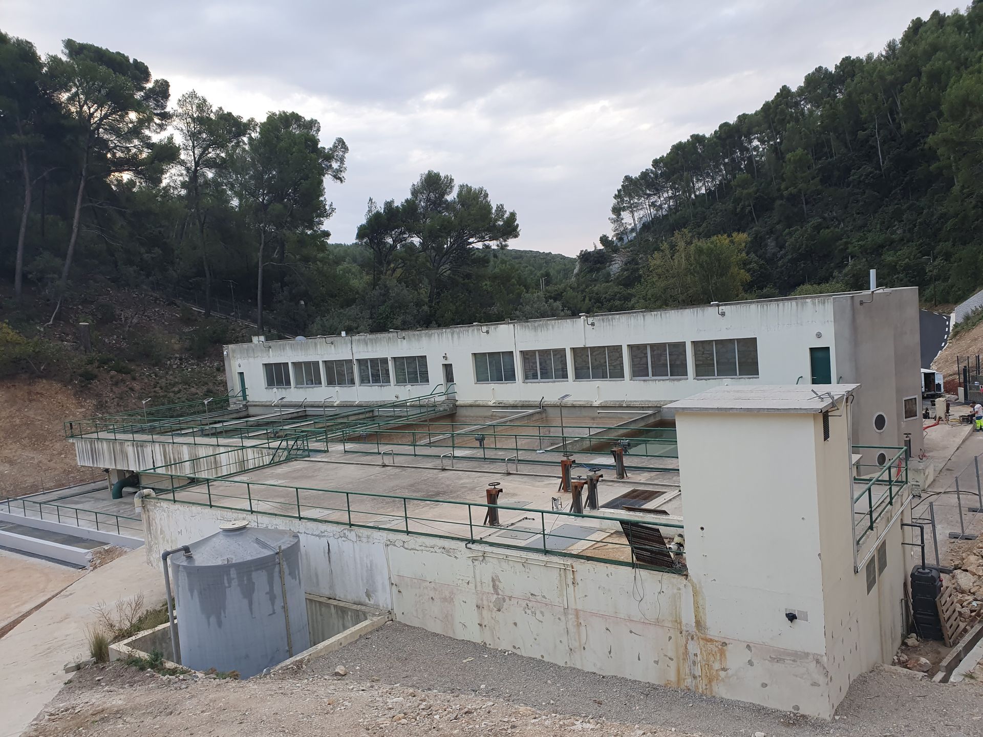 Bâtiment industriel blanc en cours de construction avec des tuyaux sur le toit et des arbres alentour, sous un ciel nuageux