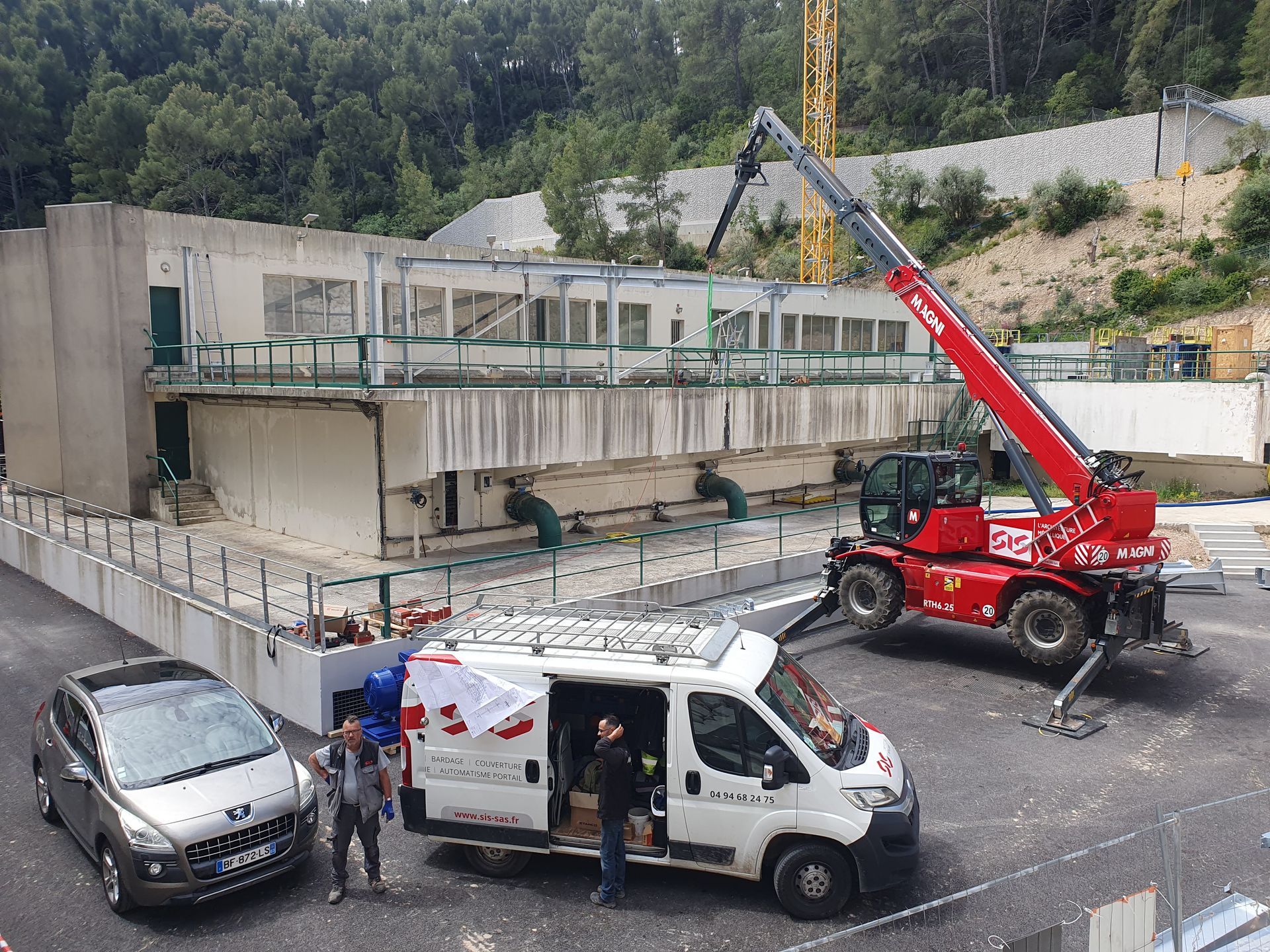 Chantier de construction avec une grue rouge soulevant des matériaux à côté d'une camionnette blanche et d'une voiture garée.