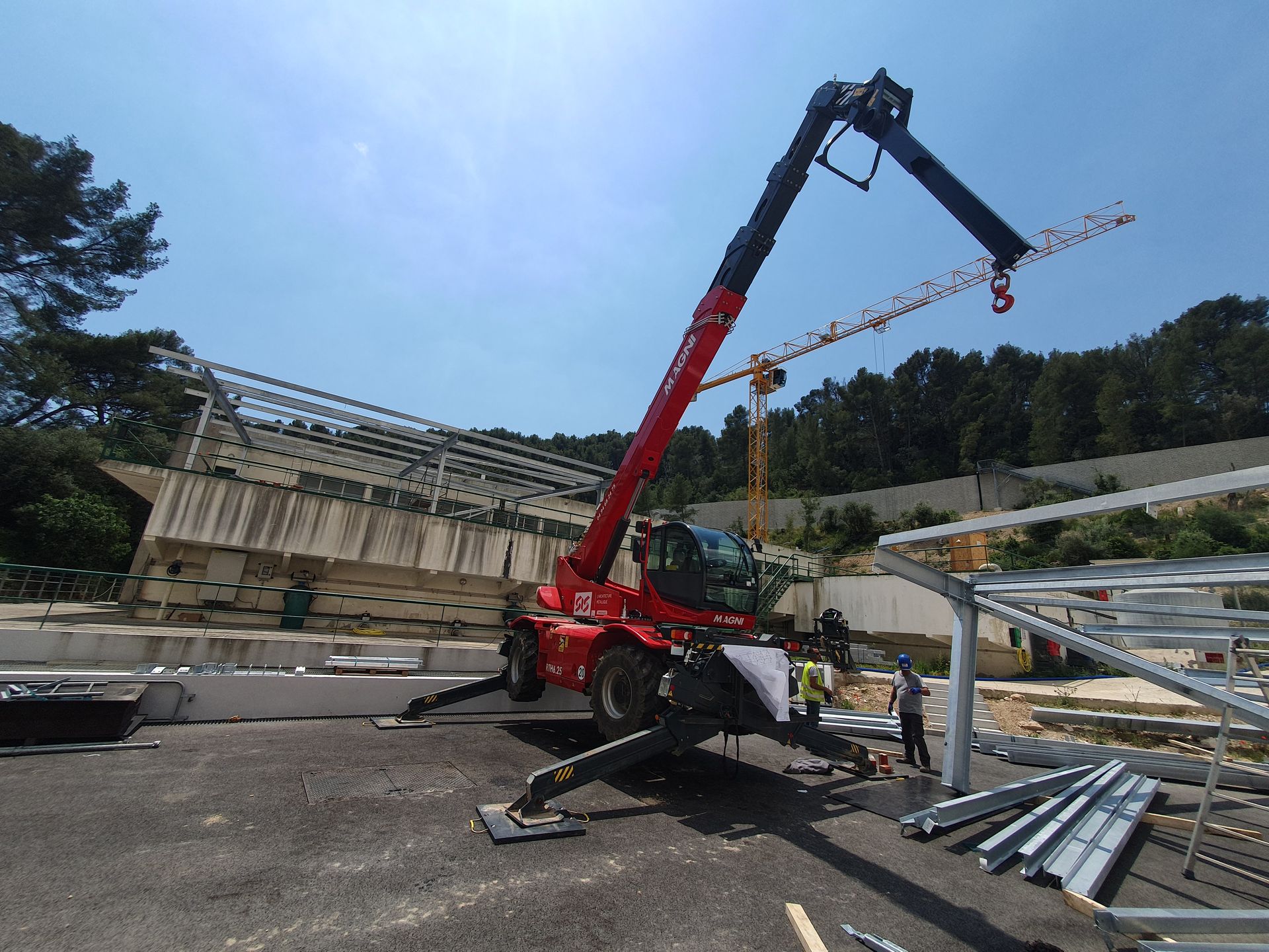 Une grue rouge soulève des poutres en acier sur un chantier en plein air, en présence d’ouvriers à proximité.