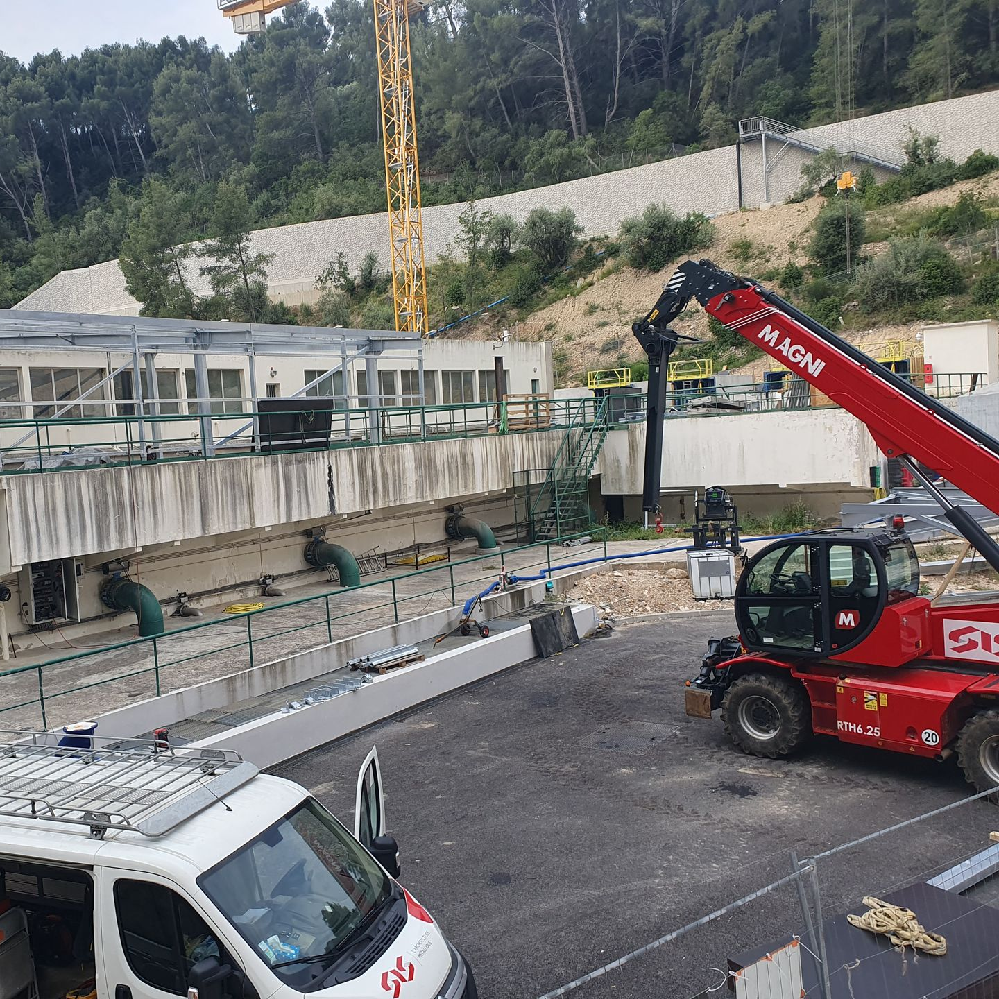 Chantier de construction avec une grue rouge soulevant des débris près d'un mur en béton et d'un flanc de colline.