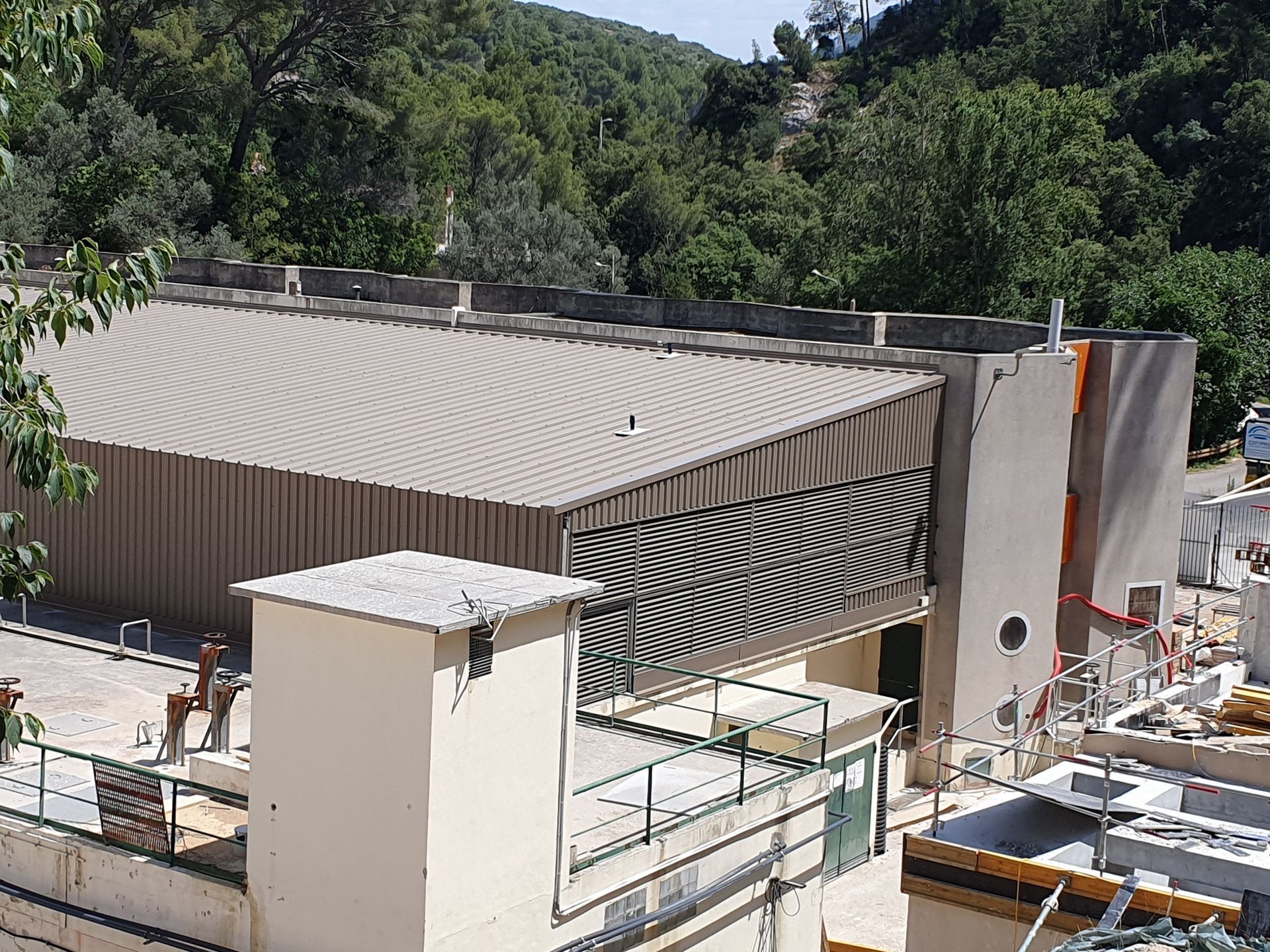 Bâtiment moderne en construction, avec toit nervuré et échafaudages, entouré d'arbres