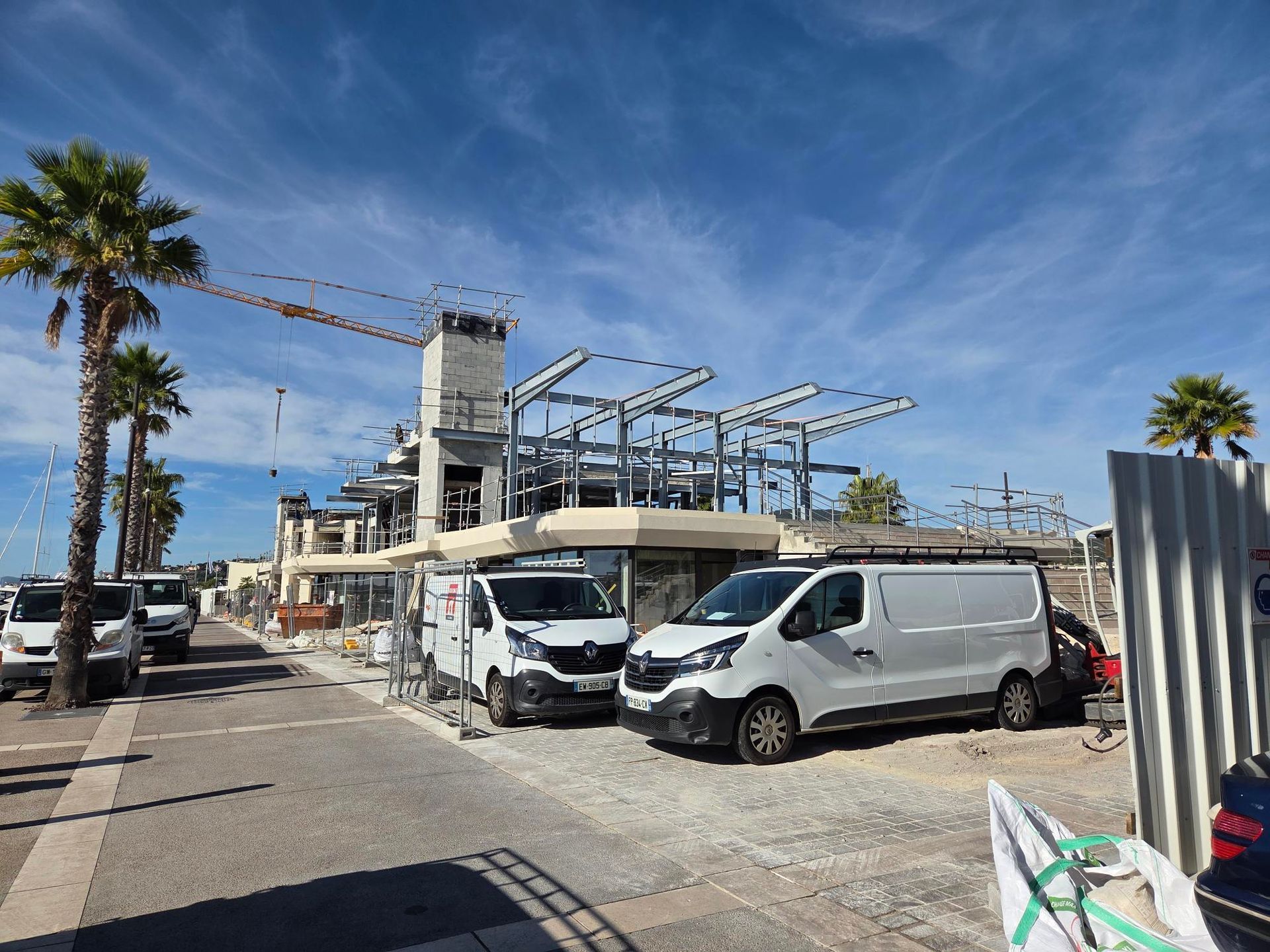 Des camionnettes garées devant un chantier de construction avec une structure métallique et des palmiers sous un ciel bleu