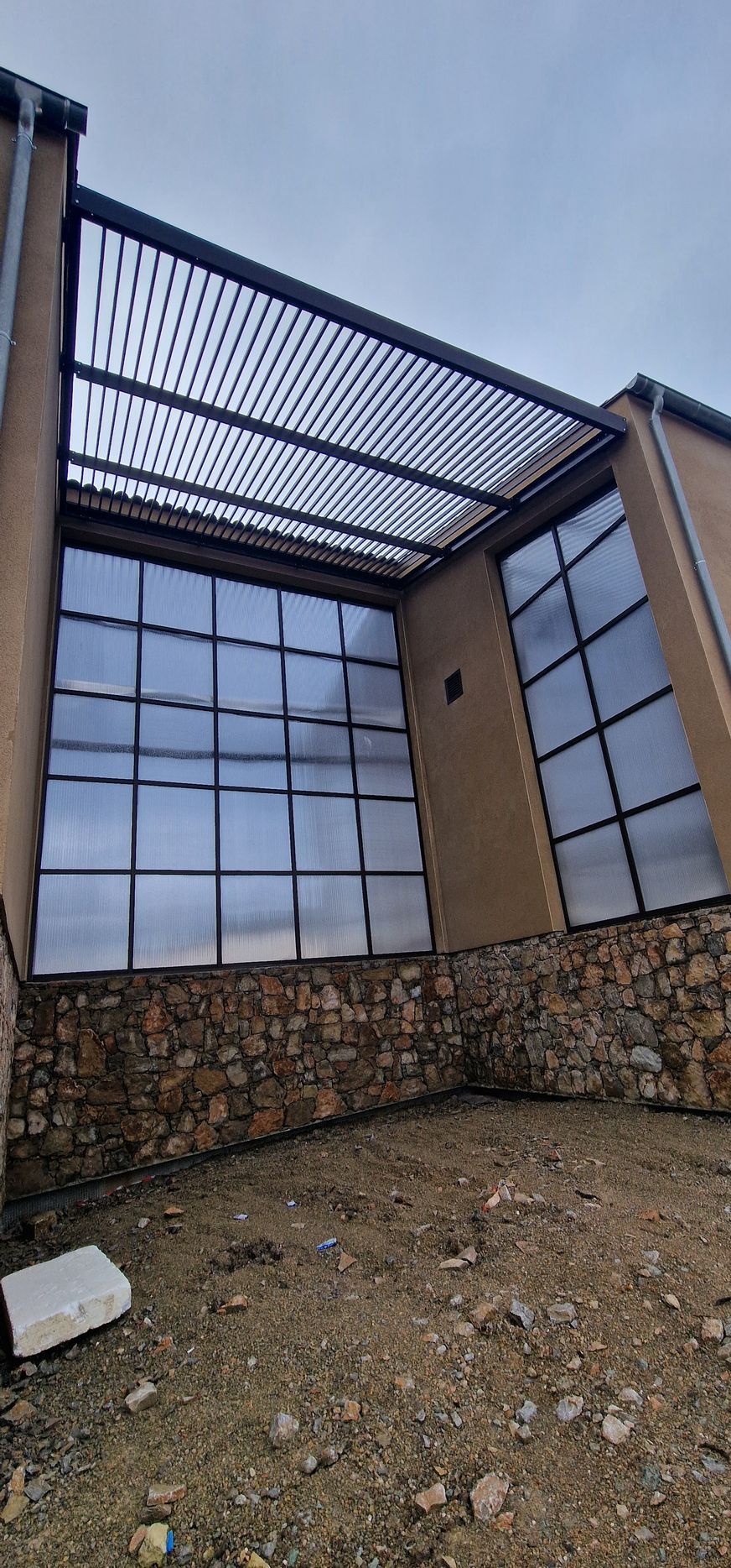 Cour intérieure d’un bâtiment moderne avec une façade vitrée, un mur en pierre et un auvent à lames métalliques.