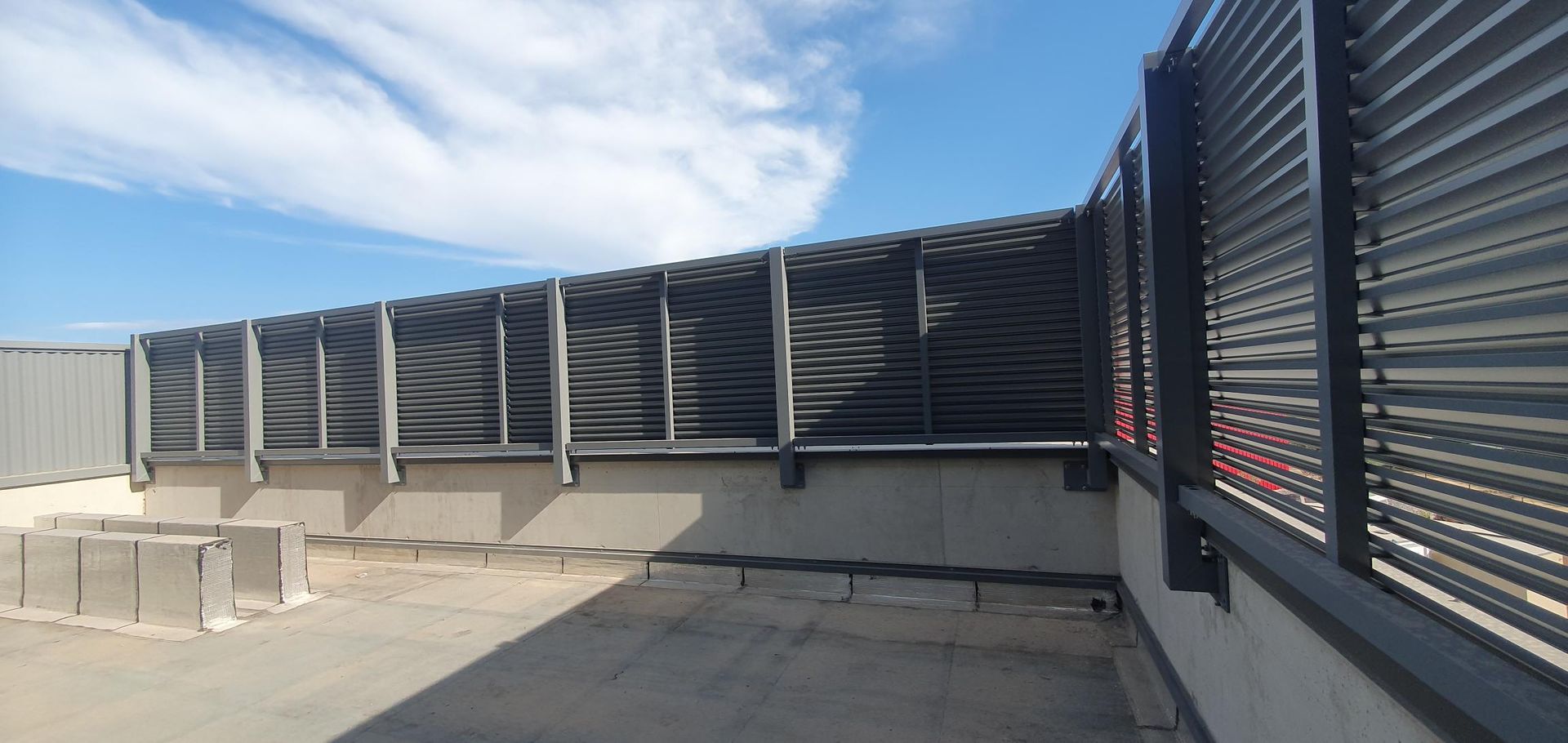 Cour industrielle extérieure avec une allée en béton bordée d'une haute clôture métallique grise, sous un ciel bleu.