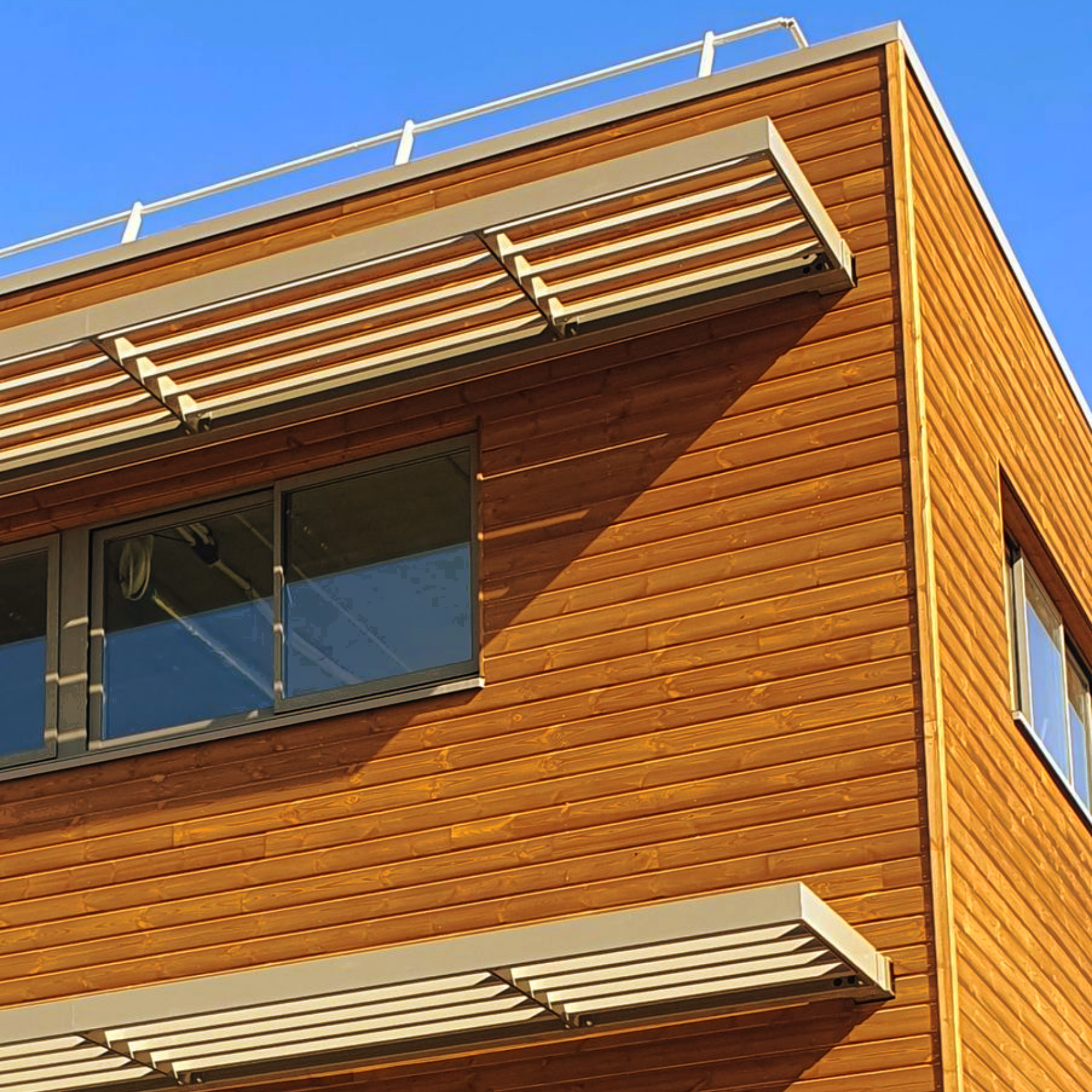 Façade moderne en bois orange d'un bâtiment, avec fenêtres et auvents déployés, se détachant sur un ciel bleu.