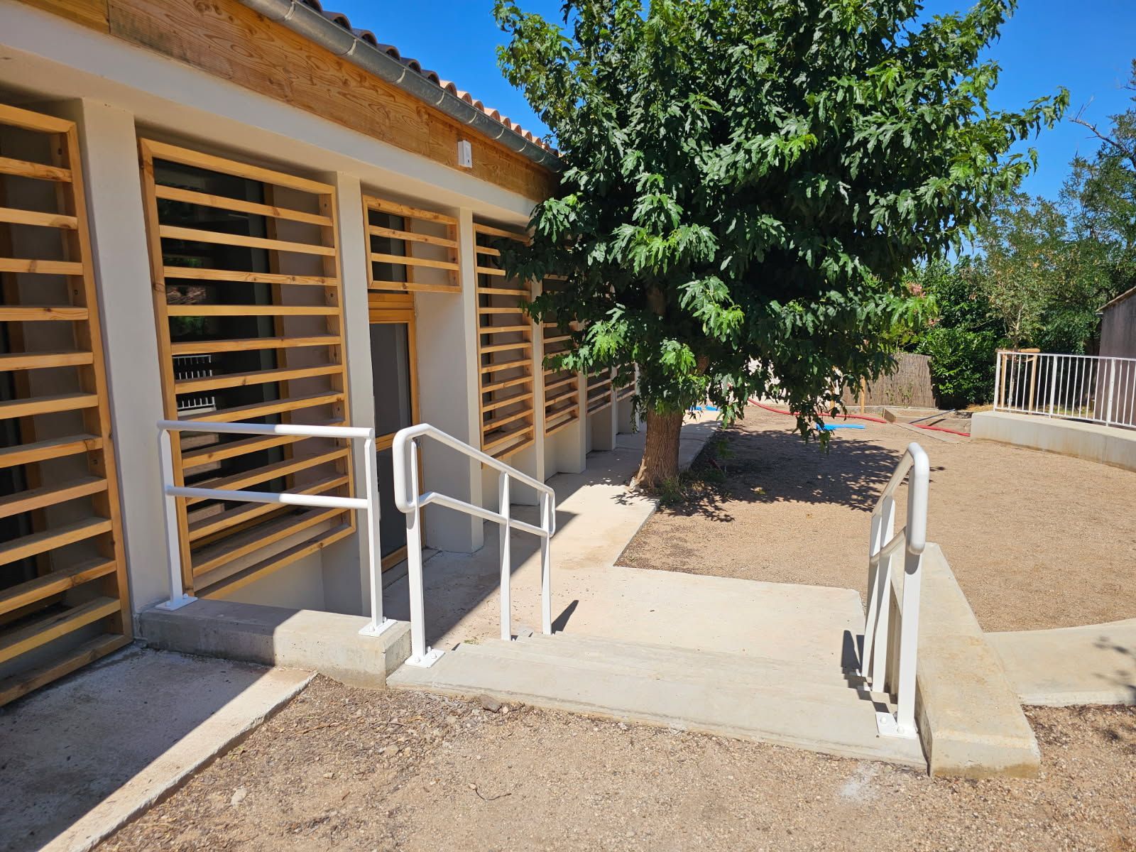 Extérieur d'un bâtiment avec une rampe d'accès pour fauteuil roulant, des balustrades et un arbre dans une cour ensoleillée.