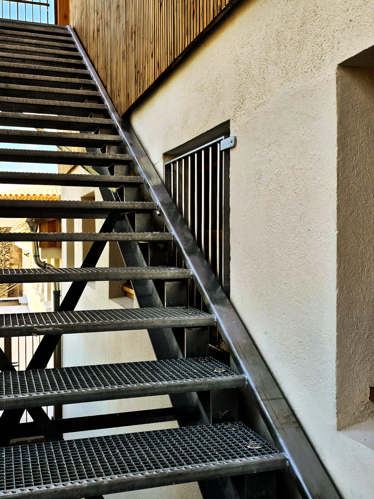 Escalier extérieur en métal, vu d'en bas, le long d'un mur beige et d'un bardage en bois.