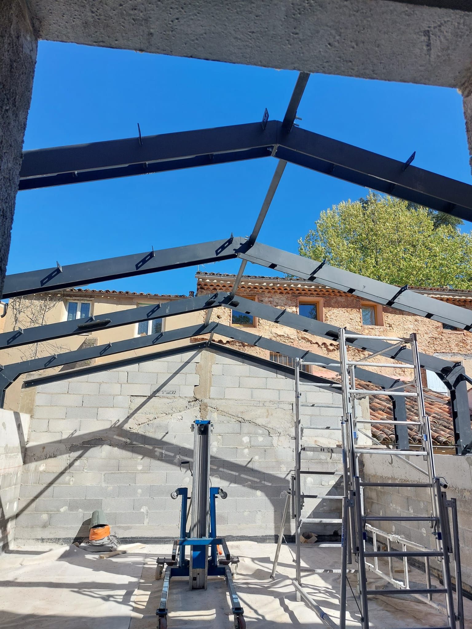 Chantier de construction avec charpente métallique, échelle et ciel bleu visible à travers le toit ouvert.