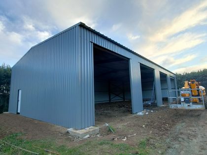 Grand hangar métallique gris en construction, avec des travées ouvertes et des engins de chantier à proximité au crépuscule