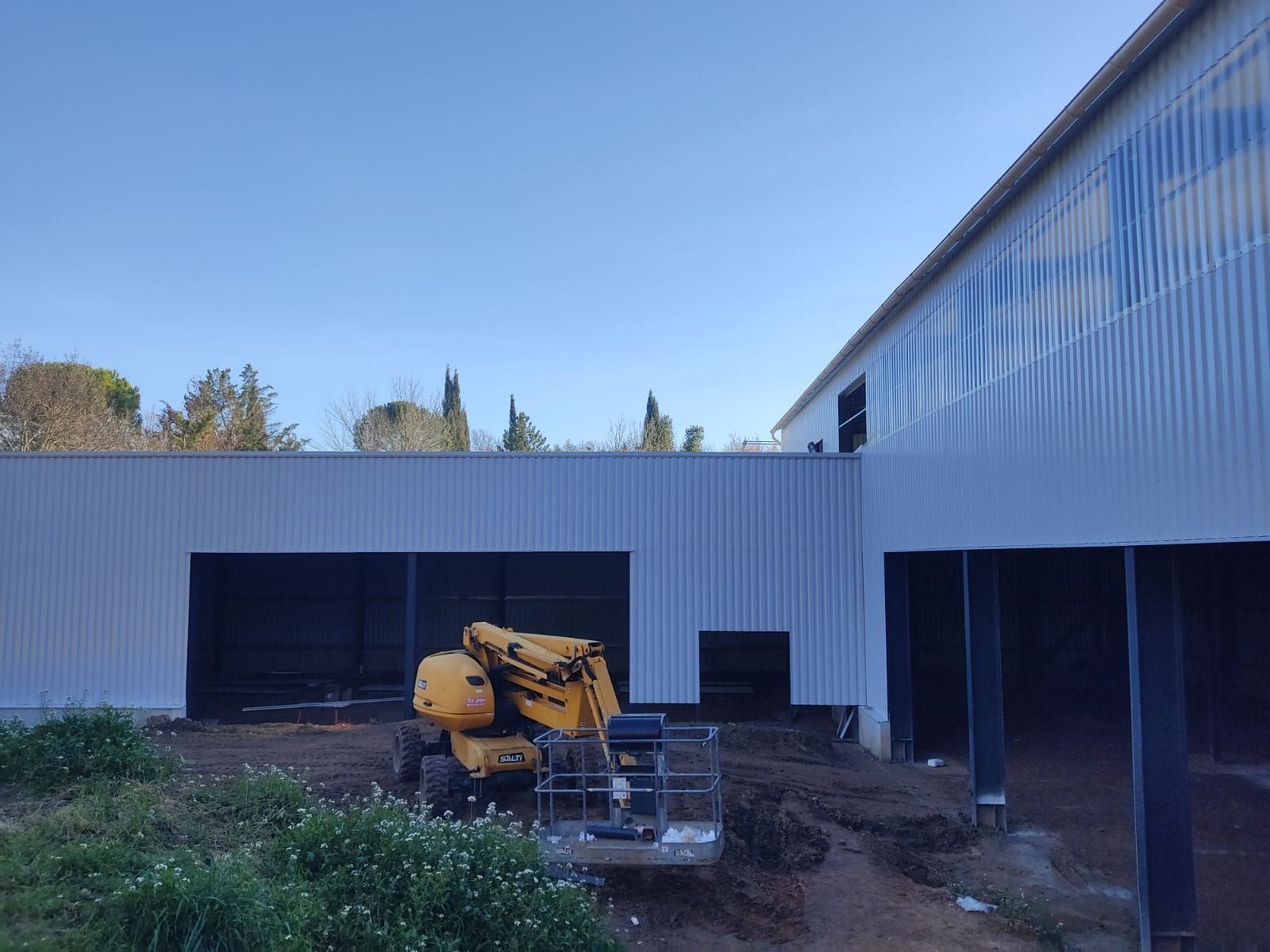 Des engins de chantier près d’un bâtiment industriel blanc, sous un ciel bleu dégagé.