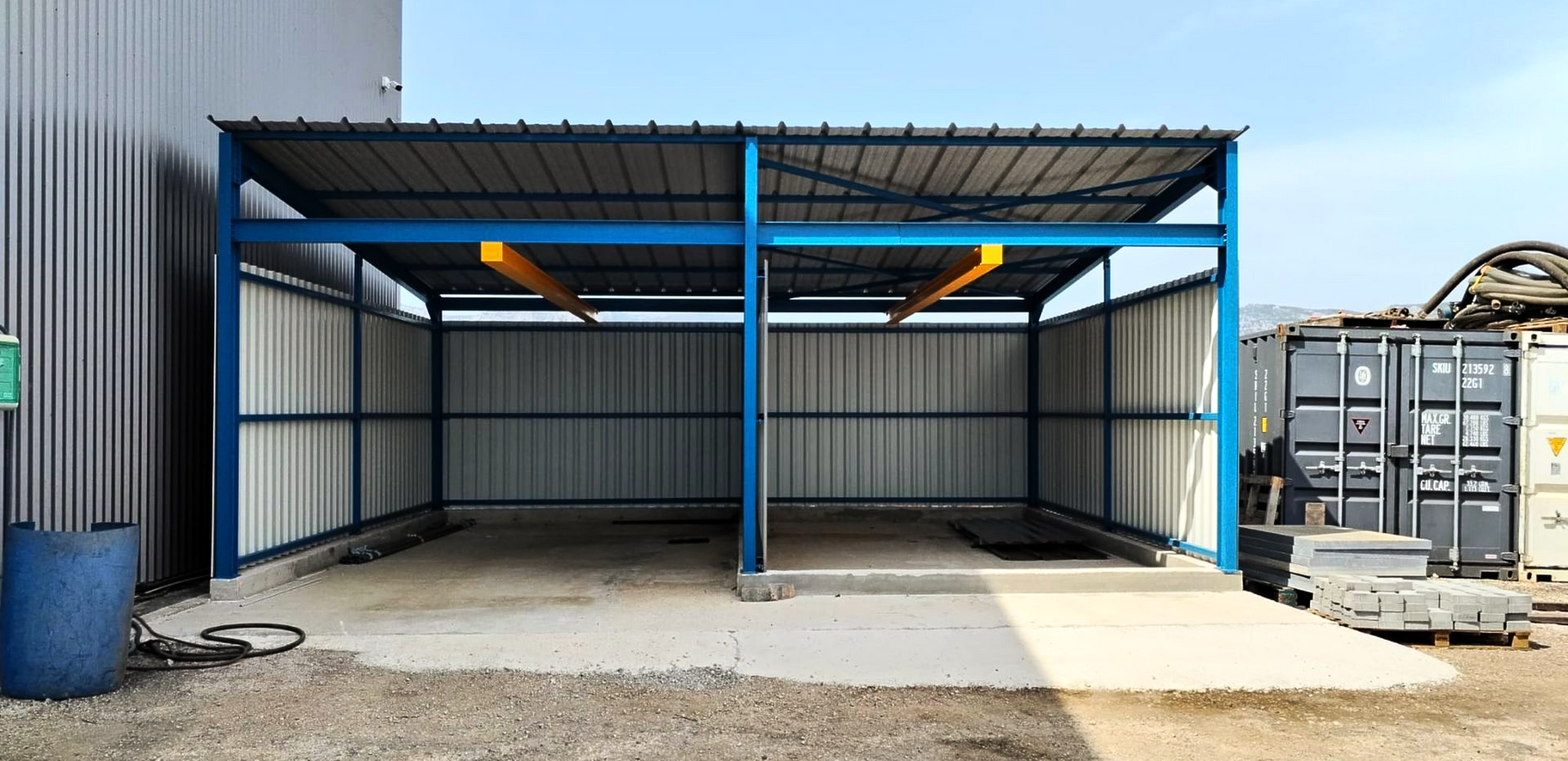 Un abri métallique se trouve à côté d'un entrepôt, avec une dalle de béton vide et des conteneurs de stockage à proximité.