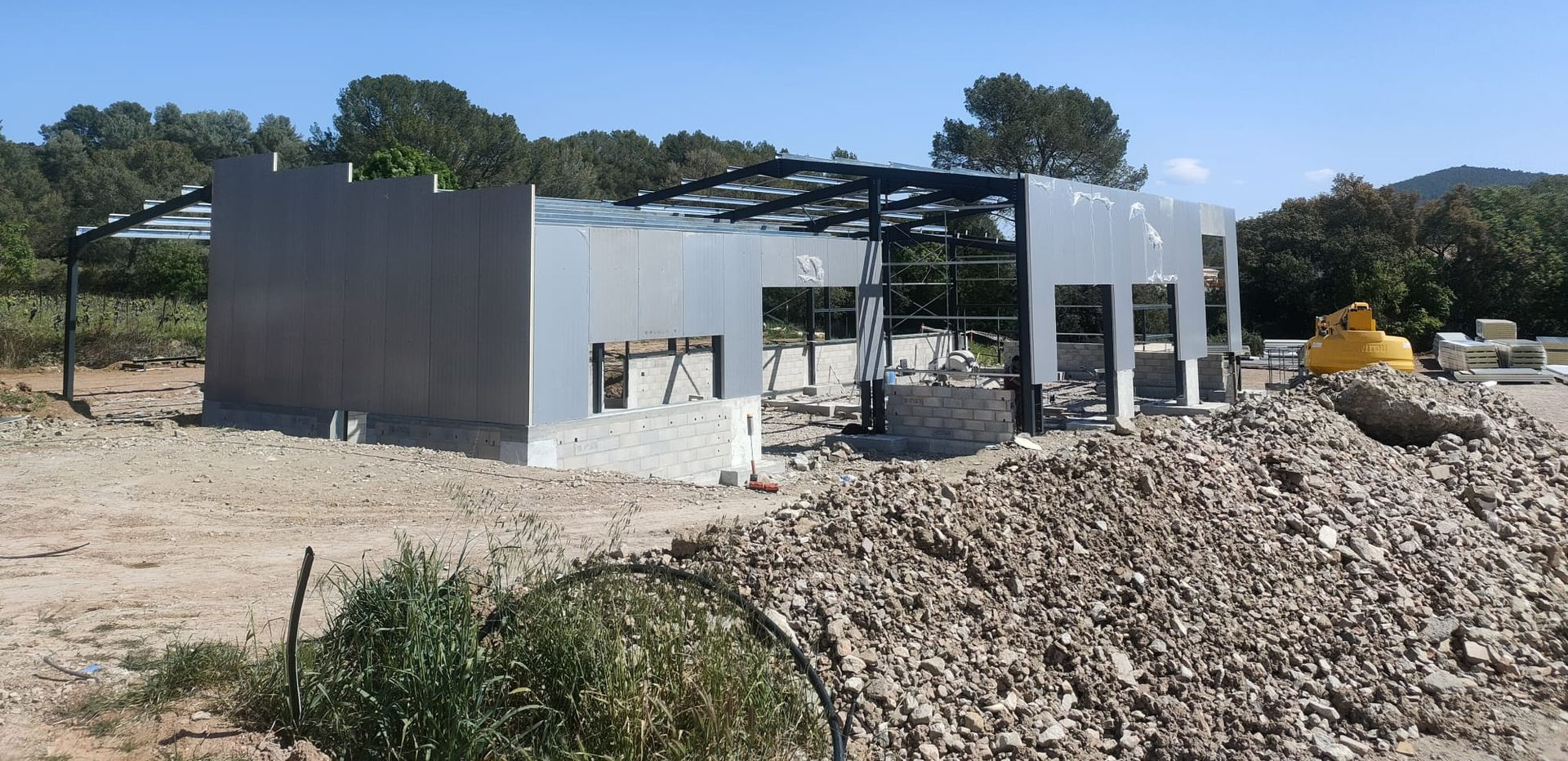 Structure métallique partiellement construite sur un chantier, entourée de gravats et d'arbres.