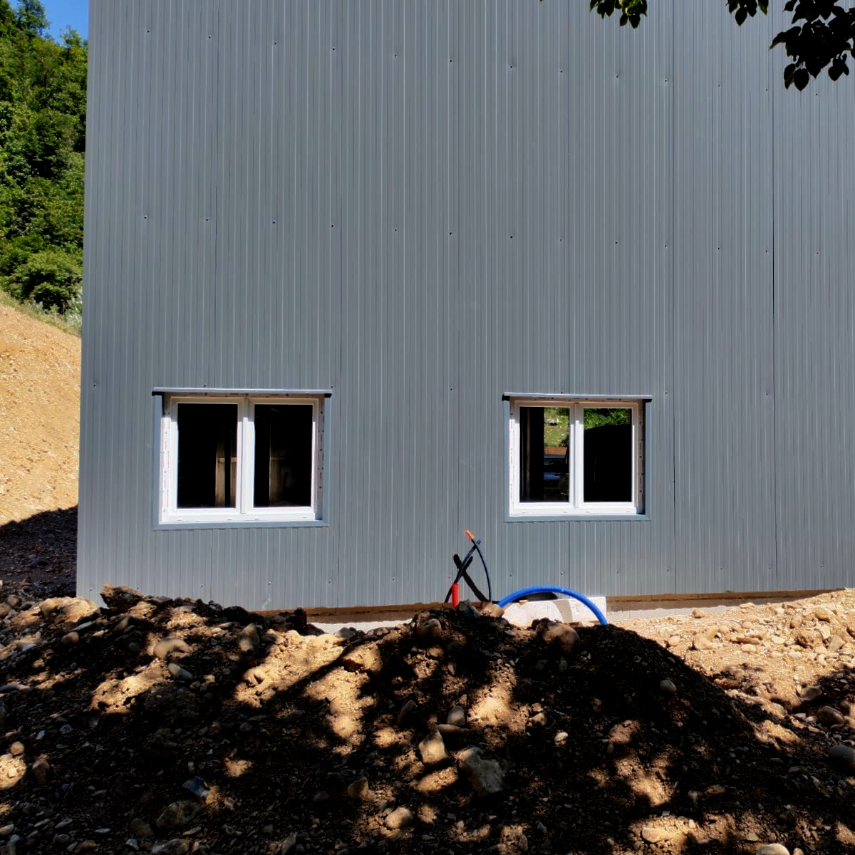 Mur d'un bâtiment en métal gris avec deux fenêtres au-dessus d'un monticule de terre et un tuyau bleu devant.